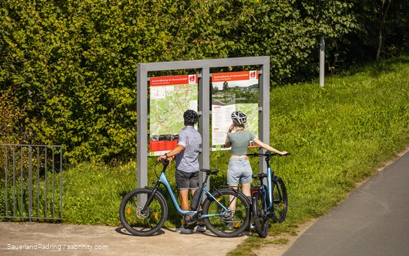Ein Radfahrerpärchen steht mit ihren Rädern vor einer Info-Tafel des SauerlandRadrings und liest.