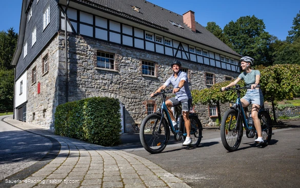 Ein Paar fährt auf E-Bikes an der Frettermühle vorbei. Im Hintergrund sieht man ein großes Gebäude aus Stein und Fachwerk.