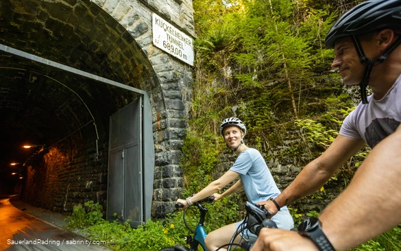 Ein Paar fährt auf E-Bikes in einen Tunnel, der als Kückelheimer Tunnel beschriftet ist.