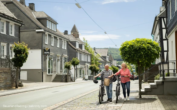 Ein älteres Paar schiebt E-Bikes entlang der Straßen von Schmallenberg. Man sieht Kopfsteinpflaster und Fachwerkhäuser mit viel Schiefer.