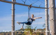 Eine Frau hängt an einem Sitzgurt an einer Metallschiene und breitet die Arme aus.