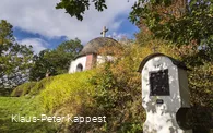 Eine Station eines Kreuzweges mit einem Teil einer Kapelle im Hintergrund umgeben von Wiesen und Bäumen.