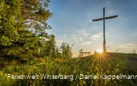 Ein großes Holzkreuz auf einem Hügel neben einer Bank vor blauem Himmel und Sonne.