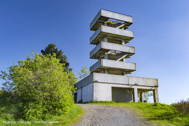Turm aus Beton mit mehreren Etagen und Treppenaufgängen vor blauem Himmel.