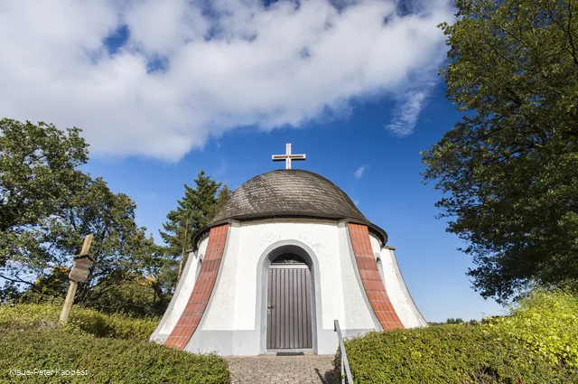 Eine Kapelle umgeben von Bäumen vor blauem Himmel.