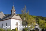 Eine Kirche vor blauem Himmel neben einem Fachwerkhaus, umgeben von Bäumen.