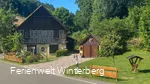 Ein altes Gebäude mit Fachwerk, davor die Einfahrt zum Grundstück und eine kleine Hütte.