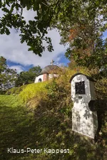 Eine Station eines Kreuzweges mit einem Teil einer Kapelle im Hintergrund umgeben von Wiesen und Bäumen.