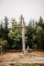 Ein großes Holzkreuz auf einer Wiese mit Wald im Hintergrund.