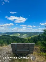 Bank auf einem Felsen mit weiter Aussicht über Wiesen und Wälder und einer Ansammlung von Häusern.