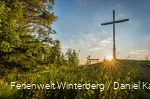 Ein großes Holzkreuz auf einem Hügel neben einer Bank vor blauem Himmel und Sonne.