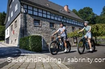 Ein Paar fährt auf E-Bikes an der Frettermühle vorbei. Im Hintergrund sieht man ein großes Gebäude aus Stein und Fachwerk.