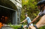 Ein Paar fährt auf E-Bikes in einen Tunnel, der als Kückelheimer Tunnel beschriftet ist.
