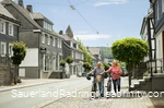 Ein älteres Paar schiebt E-Bikes entlang der Straßen von Schmallenberg. Man sieht Kopfsteinpflaster und Fachwerkhäuser mit viel Schiefer.